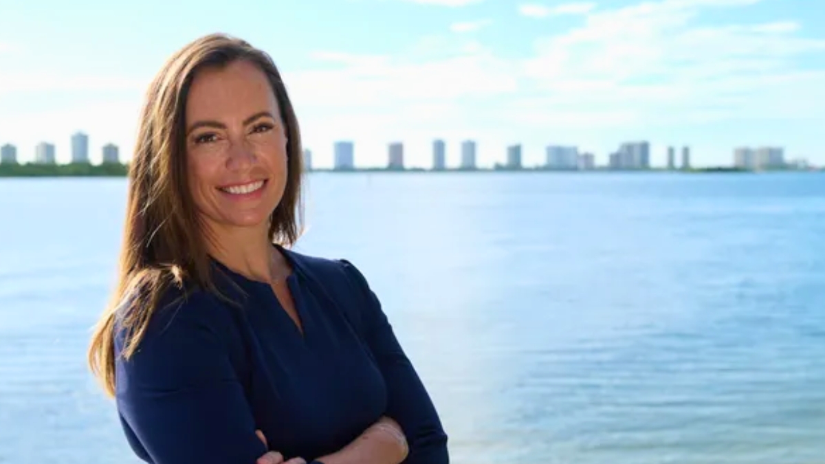 Emily Gregory smiling confidently in a navy blue dress, posed against a scenic waterfront backdrop with a city skyline on the distant horizon.