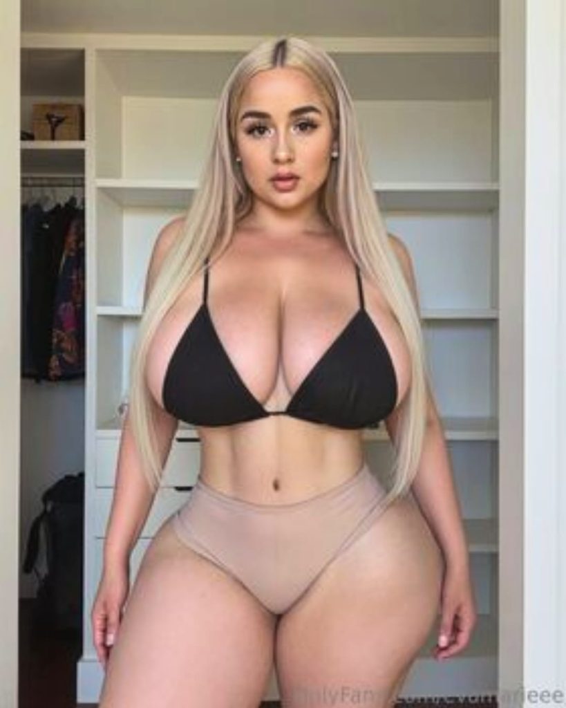 Blonde woman in black bikini top and nude bottoms posing indoors, showcasing curvy figure and long straight hair in a modern wardrobe setting