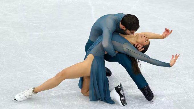 A pair of figure skaters in coordinated teal outfits perform a dramatic pose on the ice at the isu world figure skating championships 2026. The male skater is knelt on one knee, supporting the female skater as she leans back in an elegant, arched stretch with her arms gracefully extended.