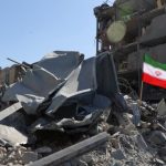 A photo of a building in partial ruins with a pile of metal and concrete rubble in the foreground. Amidst the destruction, an Iranian flag is planted on a pole, waving against the blue sky. The scene depicts the aftermath of a severe event, illustrative of the devastating consequences often seen following military actions like reported iran drone strikes in different regions.