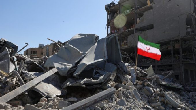 A photo of a building in partial ruins with a pile of metal and concrete rubble in the foreground. Amidst the destruction, an Iranian flag is planted on a pole, waving against the blue sky. The scene depicts the aftermath of a severe event, illustrative of the devastating consequences often seen following military actions like reported iran drone strikes in different regions.