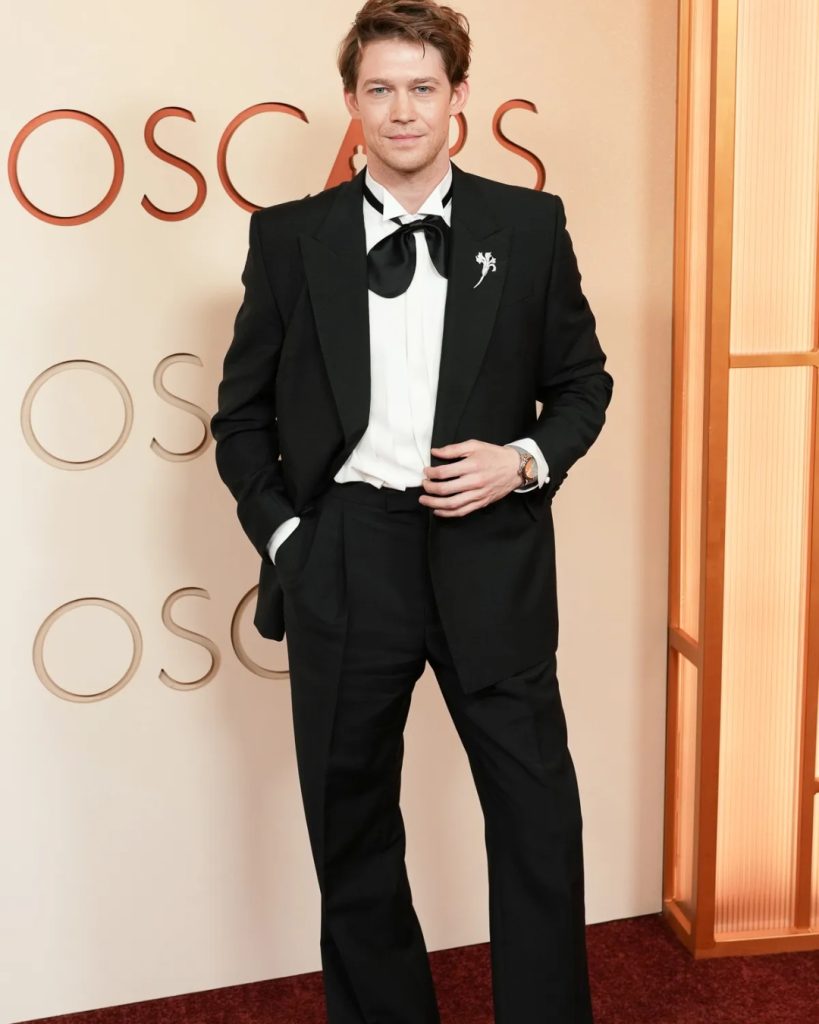 Joe Alwyn  wearing a minimalist, slim-fit black tuxedo with a crisp white shirt and a traditional black bow tie.