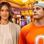 A bright composite image of Joe Burrow Jessica Alba in a Las Vegas casino setting. Jessica Alba stands on the left wearing a white top and tan blazer, looking toward the camera. Joe Burrow is on the right, wearing an orange t-shirt and a grey baseball cap worn backward, looking off-camera. Slot machines and glowing casino chandeliers are visible in the background.