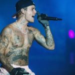 A shirtless, tattooed Justin Bieber performing on stage with a microphone, capturing the "Love Over Fame" theme of the Justin Bieber Birthday.