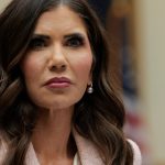 A professional close-up of Kristi Noem, often associated with the Kristi Noem Corey Lewandowski news cycle, looking slightly off-camera with a serious expression. She is wearing a light pink textured blazer, white blouse, and a single pearl drop earring while speaking at a microphone.
