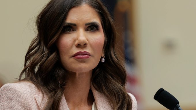 A professional close-up of Kristi Noem, often associated with the Kristi Noem Corey Lewandowski news cycle, looking slightly off-camera with a serious expression. She is wearing a light pink textured blazer, white blouse, and a single pearl drop earring while speaking at a microphone.