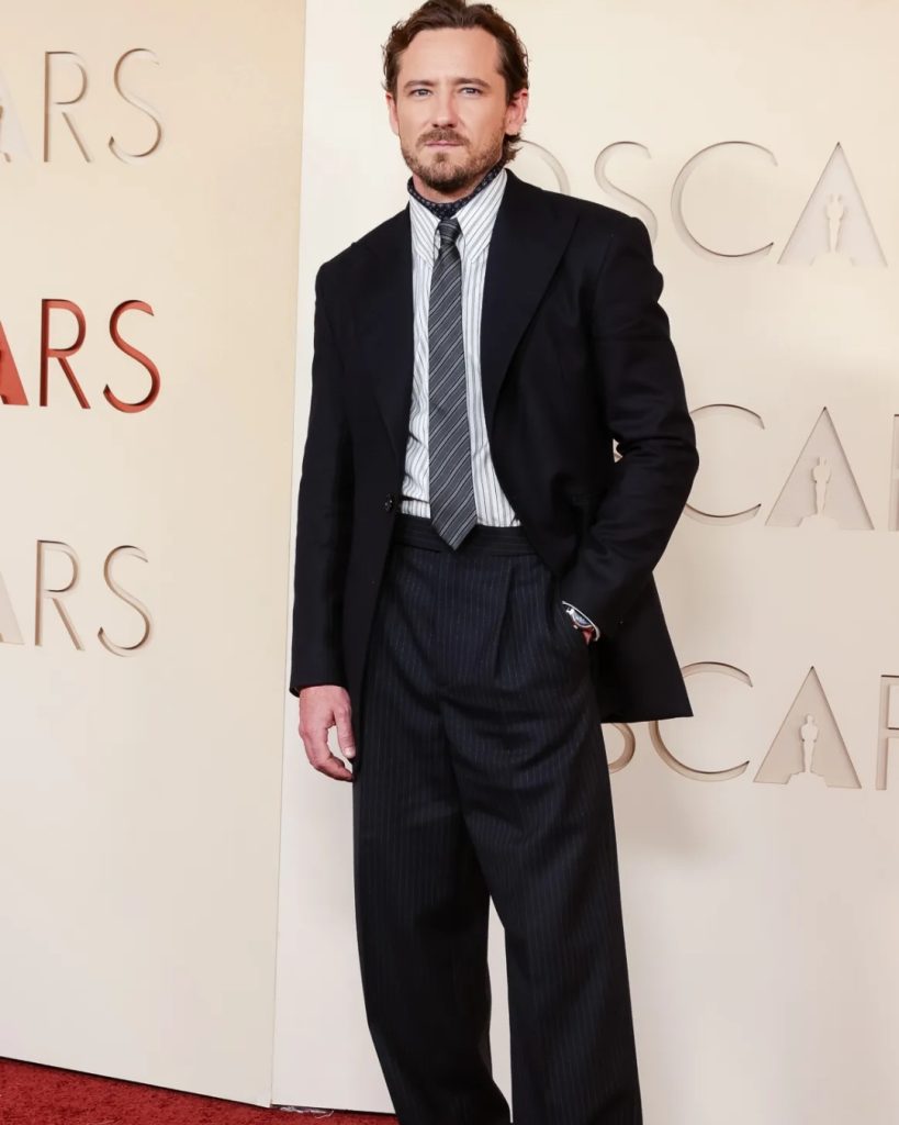 Lewis Pullman walking the Oscars Red Carpet 2026 in a midnight blue double-breasted tuxedo with black peak lapels.