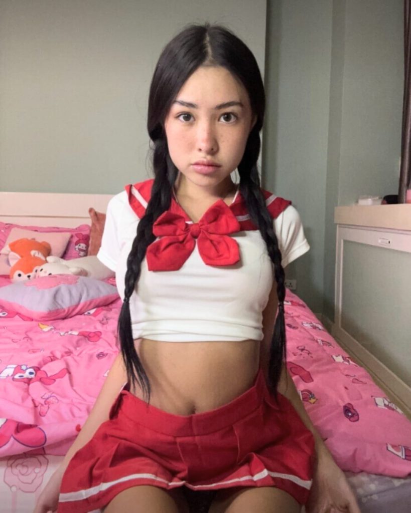 Asian model, Lil Miaa poses with braided hair wearing red and white sailor-style outfit sitting on pink themed bed.