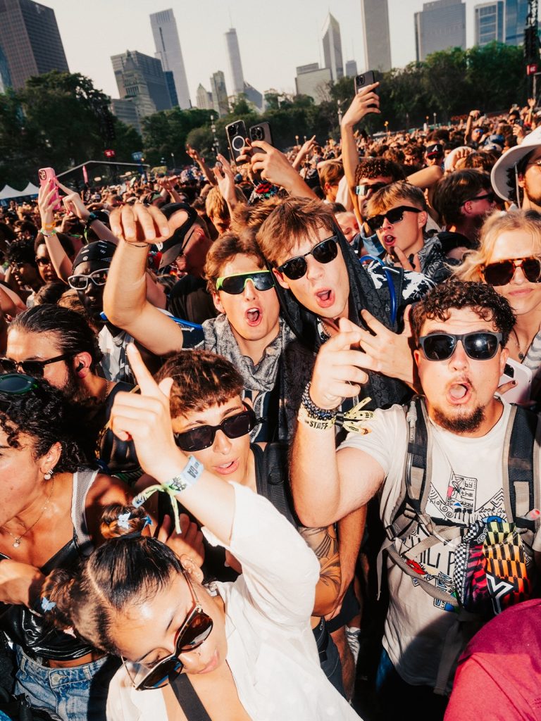 A close-up shot of energetic music fans wearing sunglasses and festival gear, cheering and posing for the camera during Lollapalooza 2026 in Chicago.