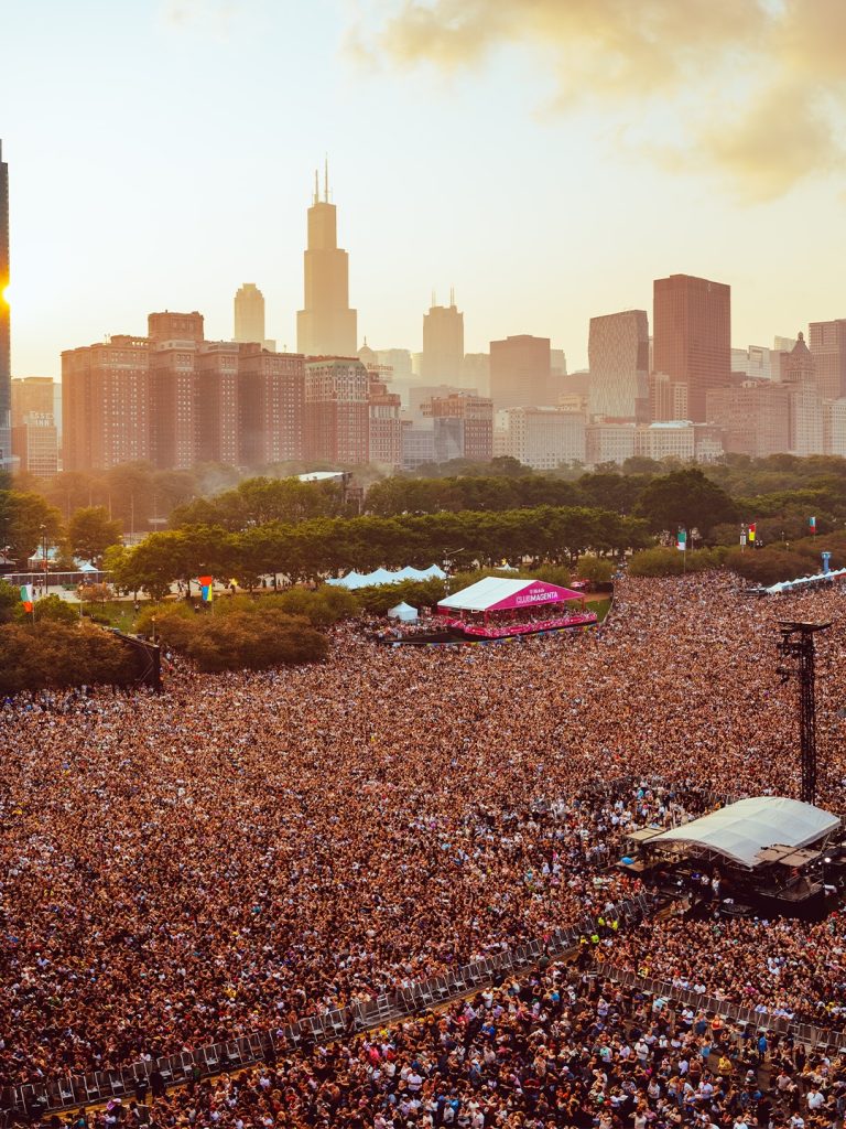 An elevated wide-angle view of a massive crowd at Lollapalooza in Grant Park, Chicago, with the city skyline and Willis Tower in the background under a sunset sky.