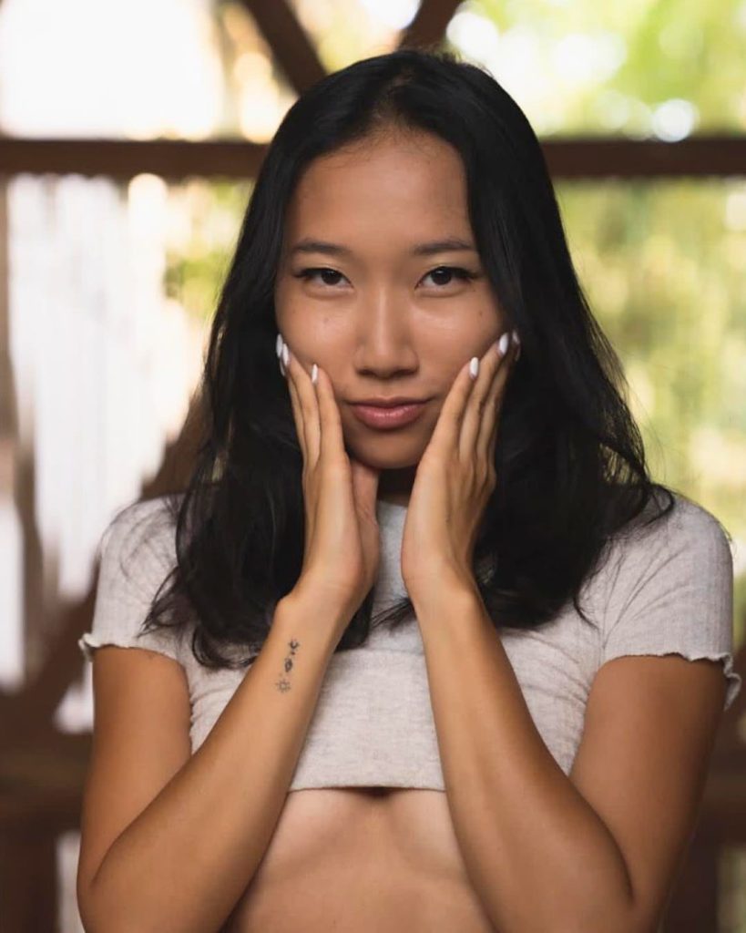 Young woman with long dark hair stands indoors, gently cupping her face with her hands. She wears a light crop top, exuding a calm, contemplative mood.