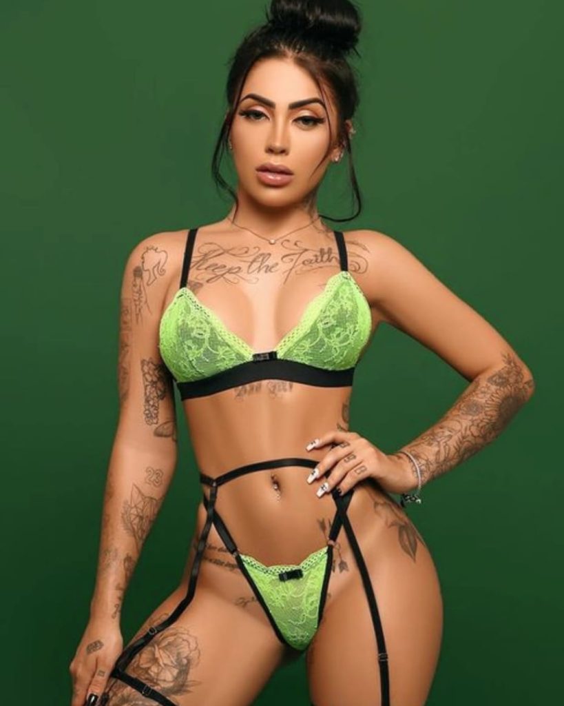 Woman in green lace lingerie poses confidently against a green background. Tattoos cover her arms and torso, creating a bold, striking look.