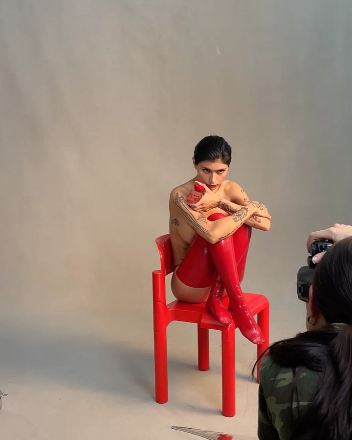 A professional studio photograph of Mia Khalifa posing on a bright red chair, wearing high-shine red boots and holding a red fragrance bottle. This artistic shot represents the high-end brand deals impacting the Mia Khalifa Net Worth.