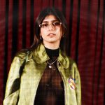 A portrait of Mia Khalifa wearing a sheer black top under a metallic green jacket with a red striped background. This stylish look highlights her fashion evolution which has contributed to the growing Mia Khalifa Net Worth.