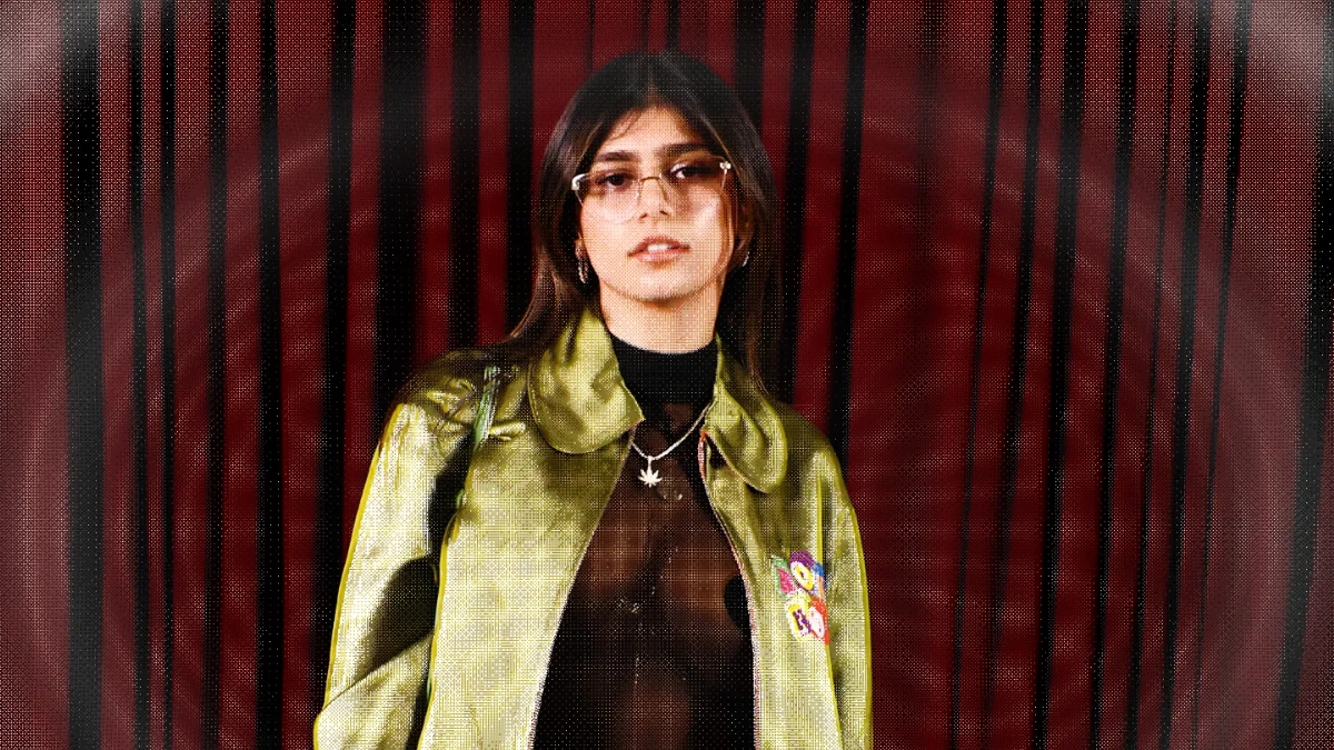 A portrait of Mia Khalifa wearing a sheer black top under a metallic green jacket with a red striped background. This stylish look highlights her fashion evolution which has contributed to the growing Mia Khalifa Net Worth.