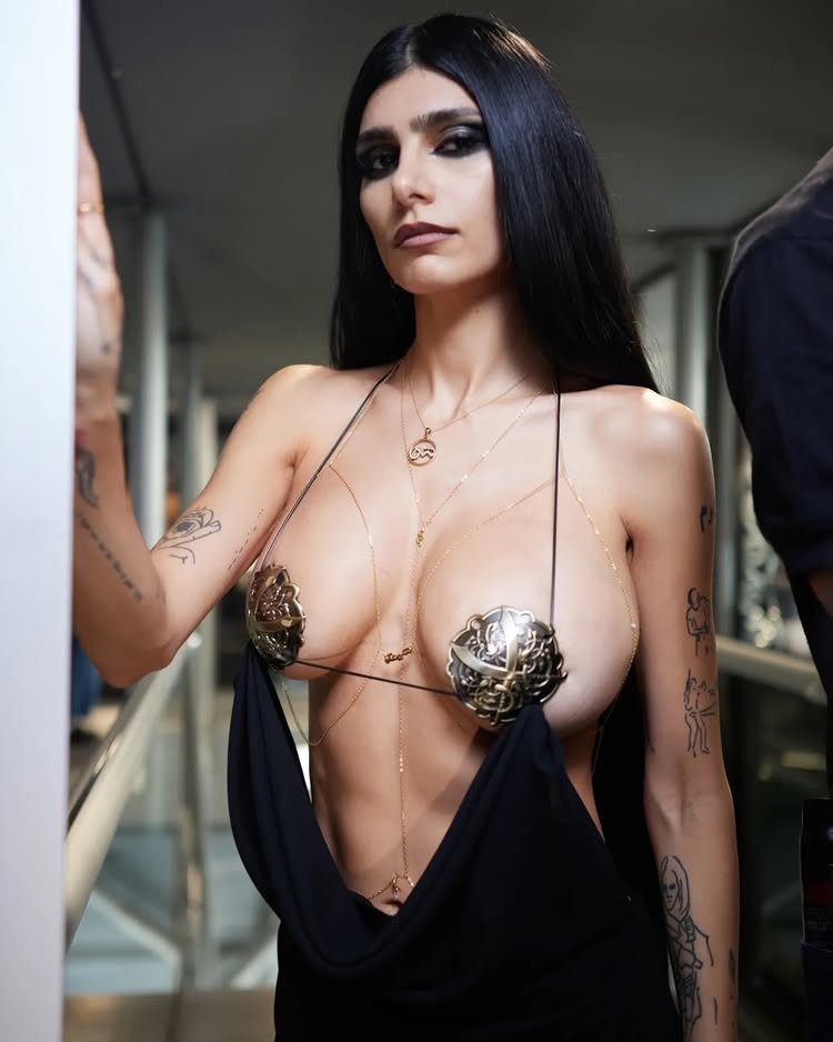 A full-length shot of Mia Khalifa at Paris Fashion Week standing against a white wall. She is wearing a high-fashion, revealing black draped gown accessorized with intricate gold filigree breastplates and thin gold necklaces. Her arms are visible, showcasing several minimalist tattoos.