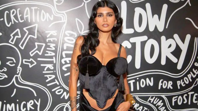 Mia Khalifa posing in front of a "Slow Factory" black-and-white graphic backdrop during a media appearance. She is wearing a black velvet corset top with distinctive tassel details and a gold cuff bracelet. Mia Khalifa News coverage often highlights her involvement with advocacy and fashion organizations.