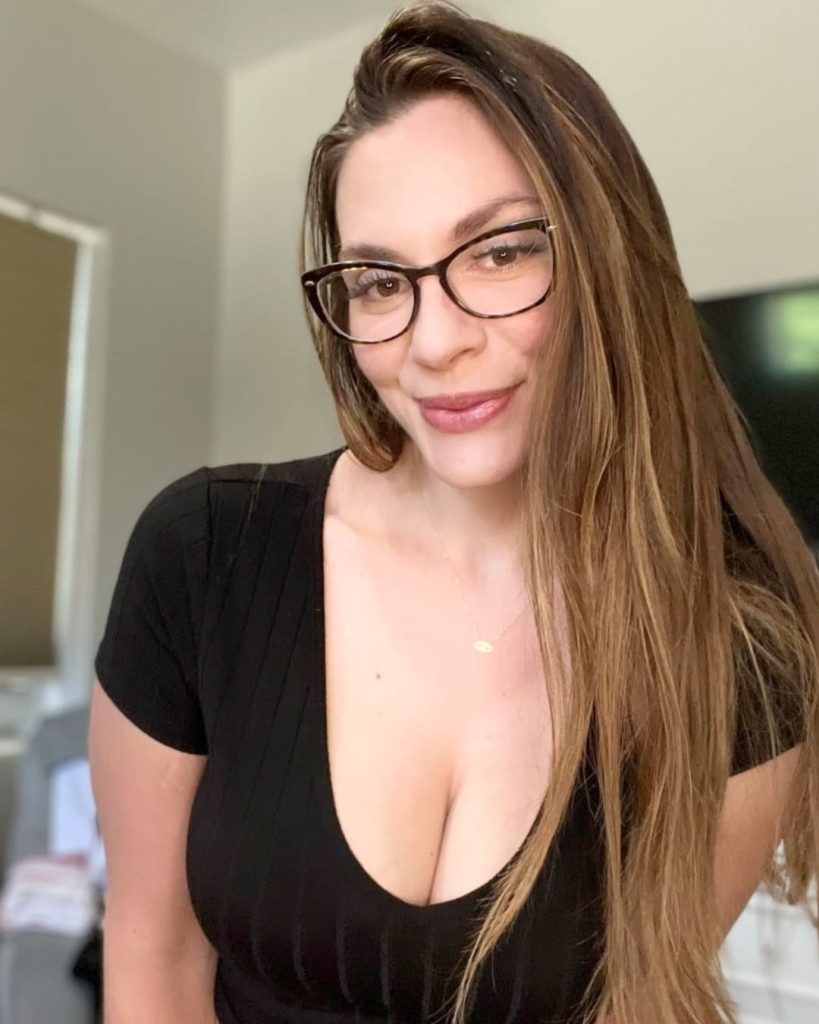 Woman with long brown hair and glasses, wearing a black top, smiles warmly. Neutral-colored room with a TV and window in the background.