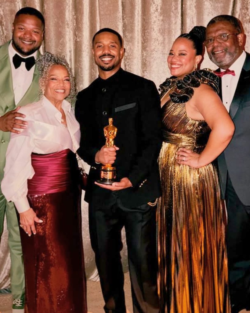 A warm group photo featuring Michael B. Jordan holding his Best Actor Oscar while posing with colleagues or family. He is dressed in a black formal suit with gold button details. The group stands against a shimmering gold curtain backdrop, with a woman in a metallic gold pleated dress to his right and a woman in a deep burgundy sequined skirt to his left.