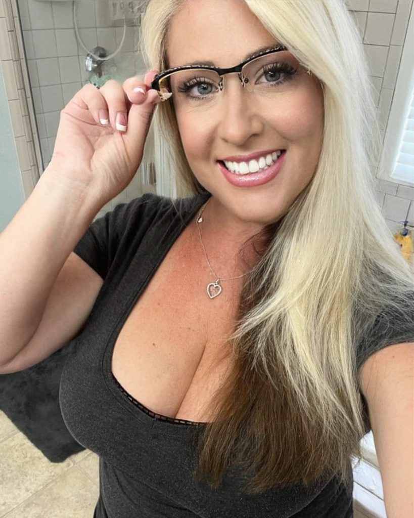 A woman with long blonde hair smiles brightly, adjusting her glasses. She's wearing a black top and heart-shaped necklace, standing in a well-lit bathroom.
