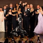 The cast and crew of the film One Battle After Another celebrate on stage after winning Best Picture at the 2026 Oscars. The group is pictured in high-fashion formal wear, with many members cheering and holding gold Oscar statues. Teyana Taylor stands at the center in a dramatic black and white feathered gown, raising her arm in victory, while other cast members, including Leonardo DiCaprio in a black tuxedo, stand nearby on the dark, polished stage.
