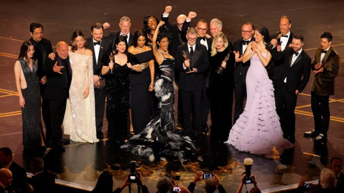 The cast and crew of the film One Battle After Another celebrate on stage after winning Best Picture at the 2026 Oscars. The group is pictured in high-fashion formal wear, with many members cheering and holding gold Oscar statues. Teyana Taylor stands at the center in a dramatic black and white feathered gown, raising her arm in victory, while other cast members, including Leonardo DiCaprio in a black tuxedo, stand nearby on the dark, polished stage.