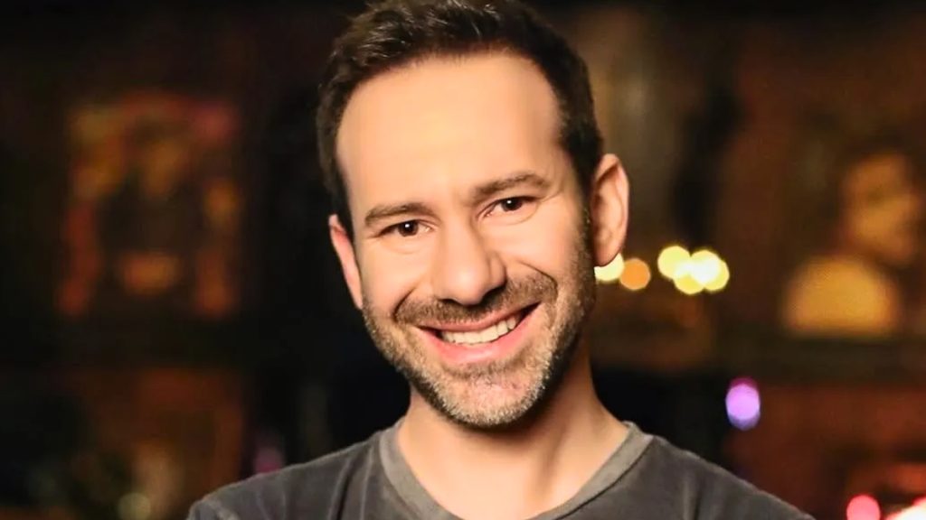 A close-up portrait of OnlyFans owner Leonid Radvinsky smiling, used in news coverage regarding the OnlyFans for sale announcement. He is wearing a grey t-shirt against a blurred, warm-toned background with bokeh lighting.