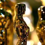 A close-up, shallow-focus shot of several gold Oscar statuettes lined up. The iconic awards gleam under warm stage lighting, with a soft, blurred "bokeh" background creating a prestigious atmosphere for the 98th Academy Awards.