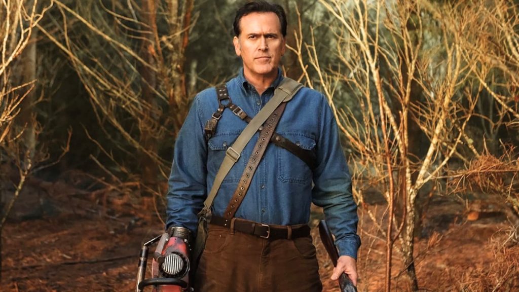 A photograph of Bruce Campbell as Ash Williams, standing in a wooded area. He is wearing a blue denim shirt with leather harnesses and a leather belt.