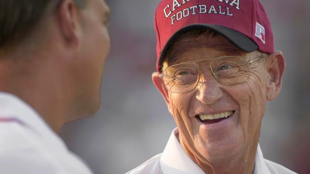 A close-up, candid photograph of legendary college football coach Lou Holtz. He is wearing a red "Carolina Football" cap and gold-rimmed glasses, smiling broadly while looking at someone just out of frame.