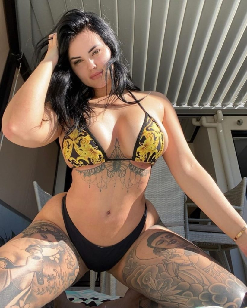 Renee Gracie posing outdoors in a yellow and black patterned bikini, showcasing her detailed torso and thigh tattoos.