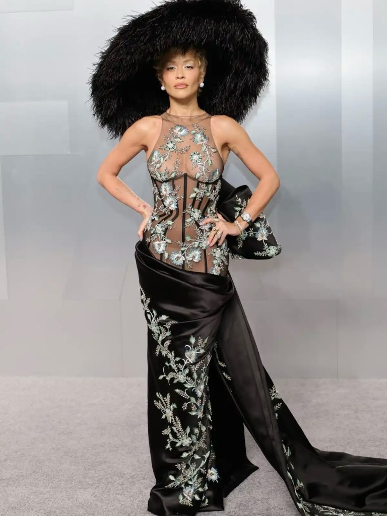 A detailed portrait of Rita Ora at the Vanity Fair Oscar Party wearing a striking, large black feathered hat and an intricate, sheer black long-sleeve gown adorned with shimmering floral embroidery.