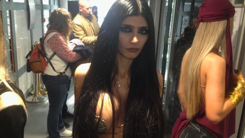 A close-up of Mia Khalifa at Paris Fashion Week, featuring her with long, dark hair and bold smokey eye makeup. She is wearing a daring black dress with metallic gold chest plates and delicate gold body chains. The background shows a crowded hallway with other attendees.