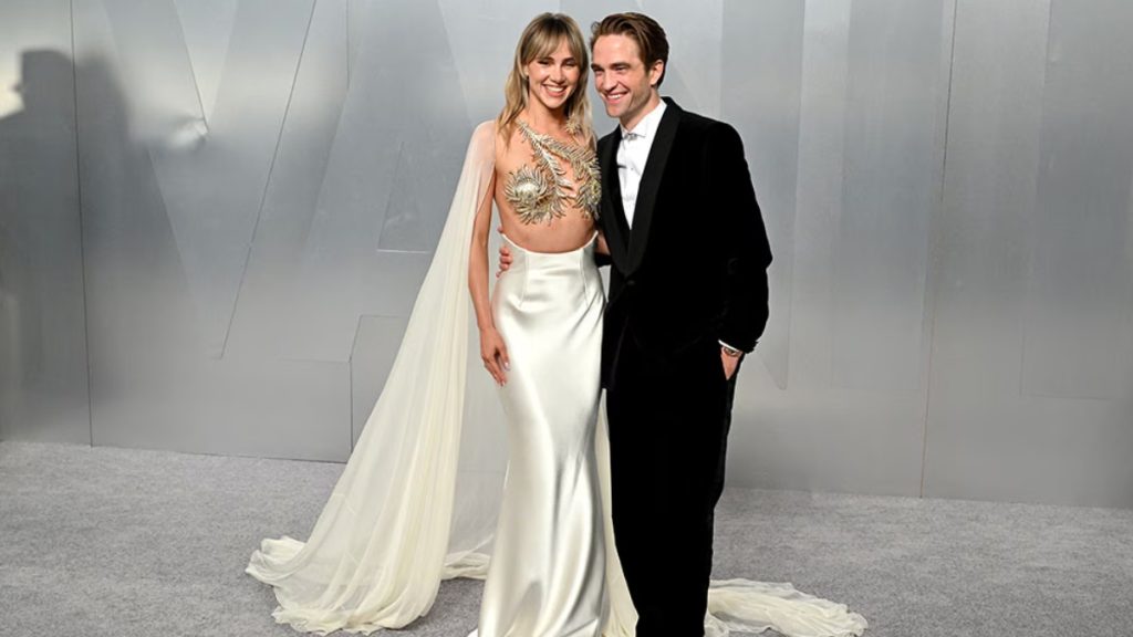 Suki Waterhouse (wearing a metallic gold anatomical bodice and cape) and Robert Pattinson on the Vanity Fair Oscar Party 2026 grey carpet.