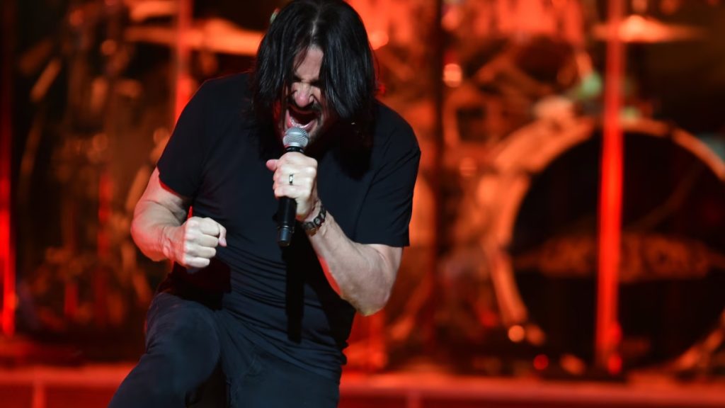A high-energy photo of musician Tommy DeCarlo performing live on stage. He is wearing a black t-shirt, leaning forward with intensity while gripping a microphone and singing. The background is filled with warm stage lights and a drum kit, capturing his legacy in classic rock.