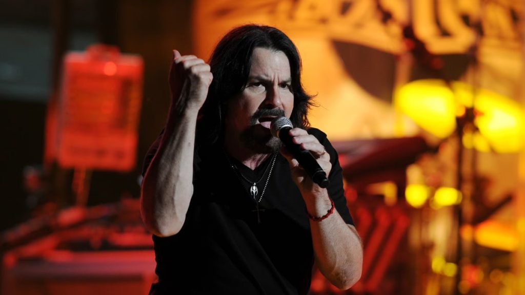 A performance photo of Tommy DeCarlo singing passionately into a microphone on stage, wearing a black t-shirt. The graphic is headlined "Tommy DeCarlo Net Worth & Career: From Ordinary to Iconic Singer," highlighting his transition to rock stardom and his financial legacy following his career with the band Boston.