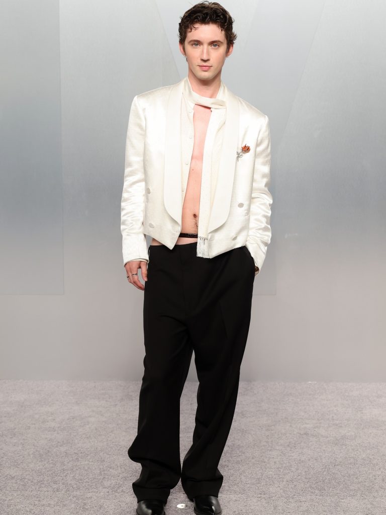 Troye Sivan wearing a daring shirtless white tuxedo look with a draped scarf on the Vanity Fair Oscar Party 2026 grey carpet.