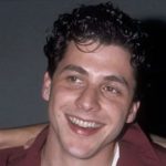 A vintage headshot of a young, smiling Corey Parker with dark curly hair, wearing a maroon shirt over a white undershirt.