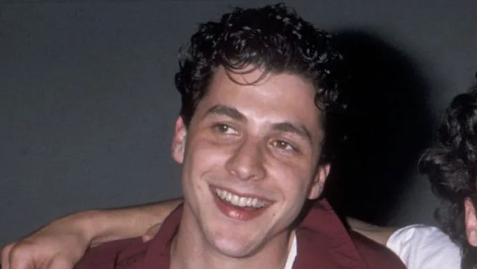 A vintage headshot of a young, smiling Corey Parker with dark curly hair, wearing a maroon shirt over a white undershirt.