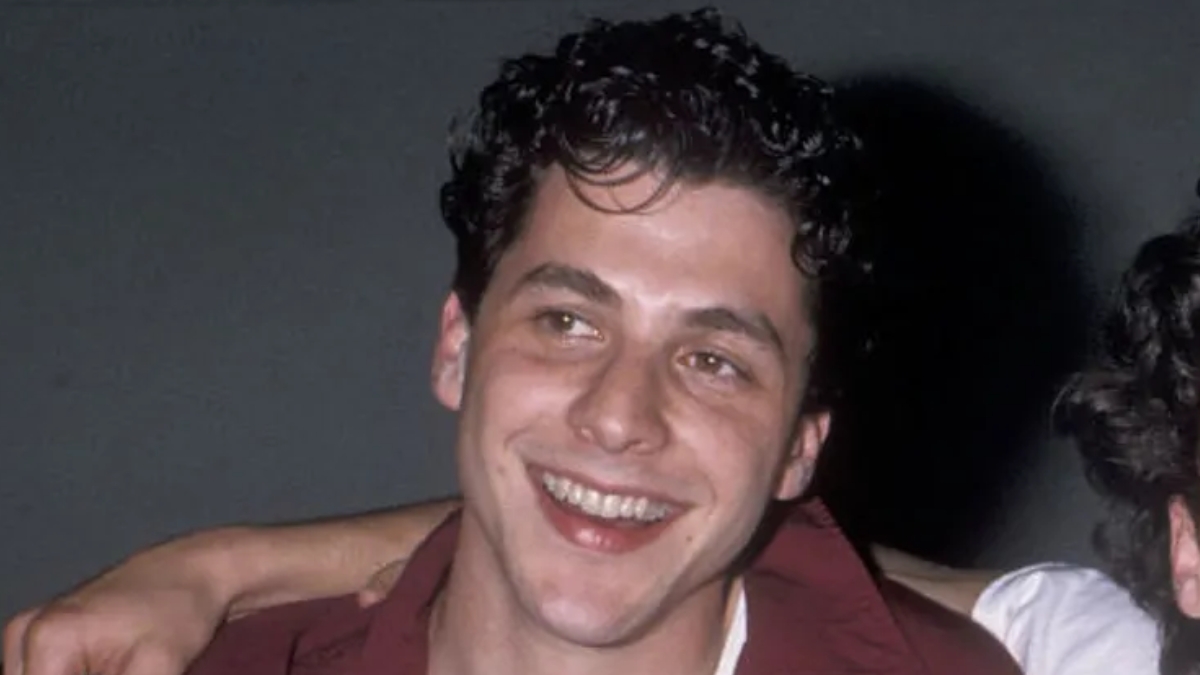 A vintage headshot of a young, smiling Corey Parker with dark curly hair, wearing a maroon shirt over a white undershirt.