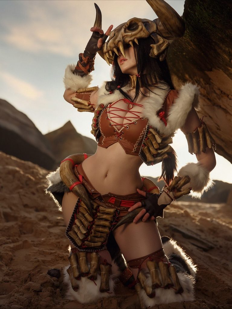 A striking cosplay portrait of Alina Becker as Nico Robin from One Piece in her "Miss All Sunday" attire. She wears a purple cowgirl hat, a lace-up purple corset top, and a matching mini skirt with white fringe. She is posed with her arms crossed in the character’s signature "Cien Fleur" ability stance against a rustic stone wall.