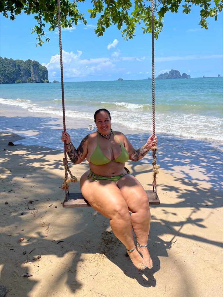 BBW OnlyFans creator April Foxx smiling on a wooden swing at a tropical beach, wearing a lime green bikini with the turquoise ocean and limestone cliffs in the background.