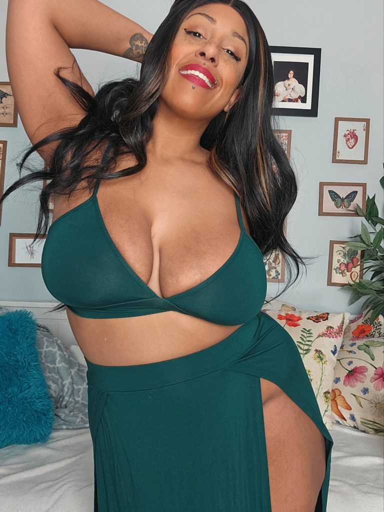 A woman with dark hair and highlights smiles while posing in a dark green two-piece outfit consisting of a bikini-style top and a matching skirt with a high leg slit. She has her hand raised to her head, revealing a small tattoo on her forearm. The wall behind her is decorated with several small, framed botanical and vintage-style illustrations.