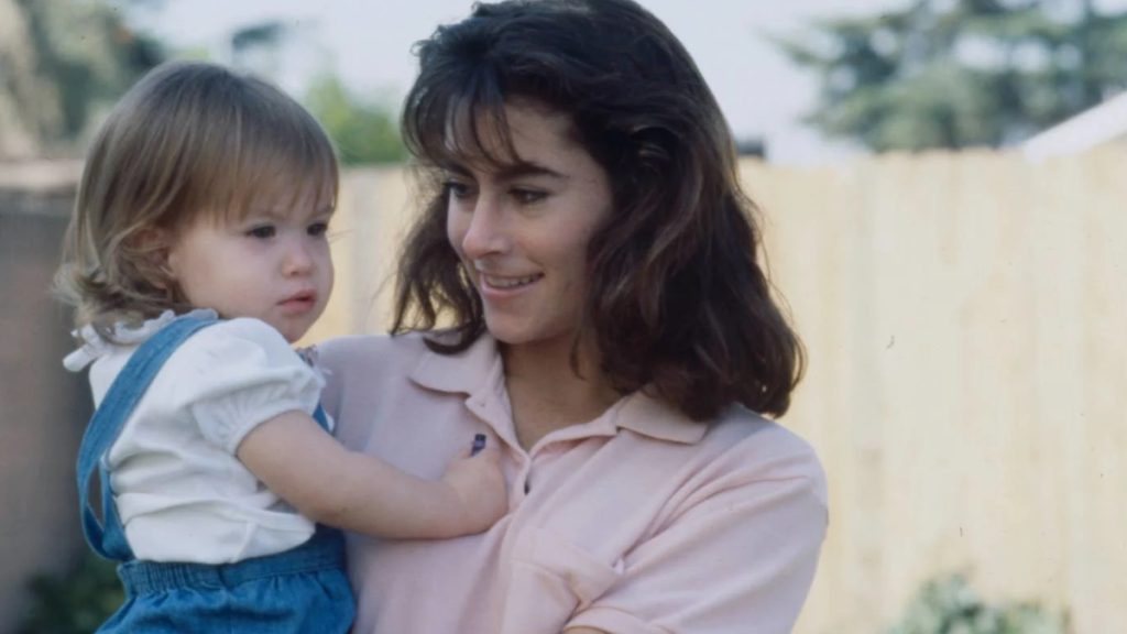 A vintage photograph of a young Jessica McClure as a toddler, being held by her mother, Reba "Cissy" McClure. Jessica is wearing denim overalls and a white ruffled shirt, looking off-camera, while her mother smiles warmly at her.