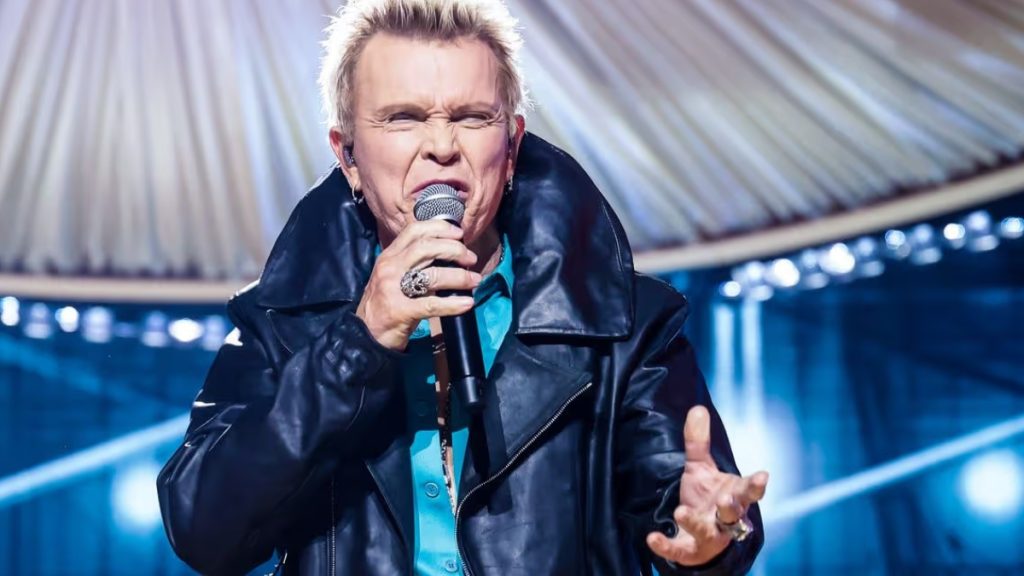 Billy Idol performing live on stage with his arms raised and fists clenched in a triumphant pose. He is biting his lip with a playful, high-energy expression. He wears an open teal shirt revealing a silver pendant necklace, topped with a black leather jacket. The background features warm stage lighting and the silhouette of a guitar headstock in the foreground.