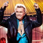 A close-up shot of Billy Idol performing on stage. He is singing intensely into a microphone, his face contorted with emotion. He sports his iconic bleached blonde spiky hair and is wearing a high-collared black leather jacket over a teal button-down shirt. A large skull ring is visible on his finger.