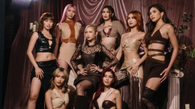A professional group shot of BINI; several members stand while others are seated, all looking away from the camera with serious expressions. The lighting is dramatic, highlighting their stylish neutral-toned outfits in a studio setting.