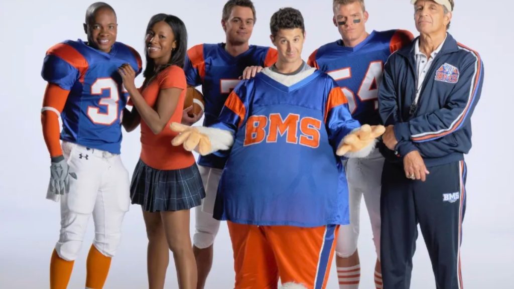An ensemble cast photo for Blue Mountain State against a white background. The group includes actors in Mountain Goats football uniforms, a cheerleader, and a coach, with a central character wearing an oversized blue "BMS" mascot jersey and furry gloves.