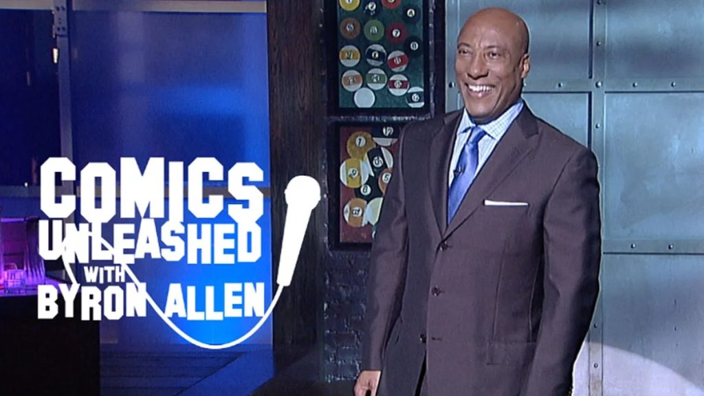 Byron Allen stands on a stage wearing a dark brown suit and a blue tie, smiling broadly. To his left, a large white graphic reads "Comics Unleashed with Byron Allen" featuring a microphone icon. The background features a dark wall adorned with two framed displays of billiard balls.