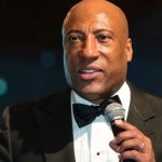 A close-up portrait of Byron Allen speaking into a microphone. He is dressed formally in a black tuxedo with a black bow tie. The background is softly blurred with teal and dark tones, highlighting his expression as he addresses an audience.